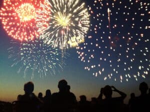A Fourth of July fireworks display exploding in the sky over a group of people. The crowd, spectators in silhouette, celebrate the Independence Day national holiday together, looking up and watching bursts of color. The pyrotechnic performance may represent New Year’s Eve, Chinese New Year, or other traditional festival celebration events.